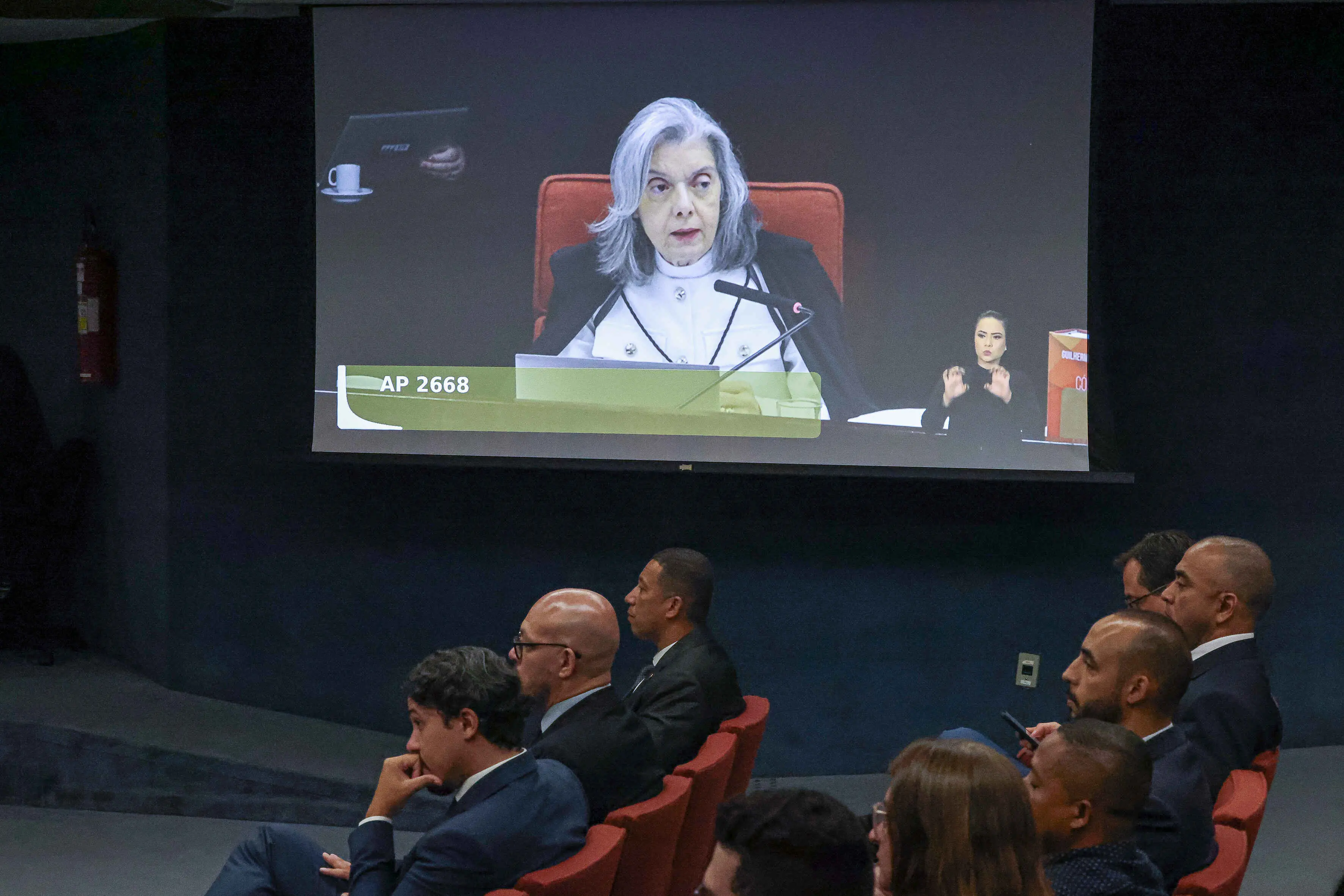 A foto mostra uma plateia assistindo, em um auditório, à transmissão de uma sessão. No telão, aparece Cármen Lúcia, uma mulher de cabelos grisalhos, vestindo toga preta sobre roupa clara, sentada diante de um microfone e um notebook, com expressão séria. No canto inferior direito da tela, há uma intérprete de Libras. Na barra inferior do vídeo, lê-se “AP 2668”. As pessoas sentadas no auditório estão voltadas para o telão, algumas usando terno.