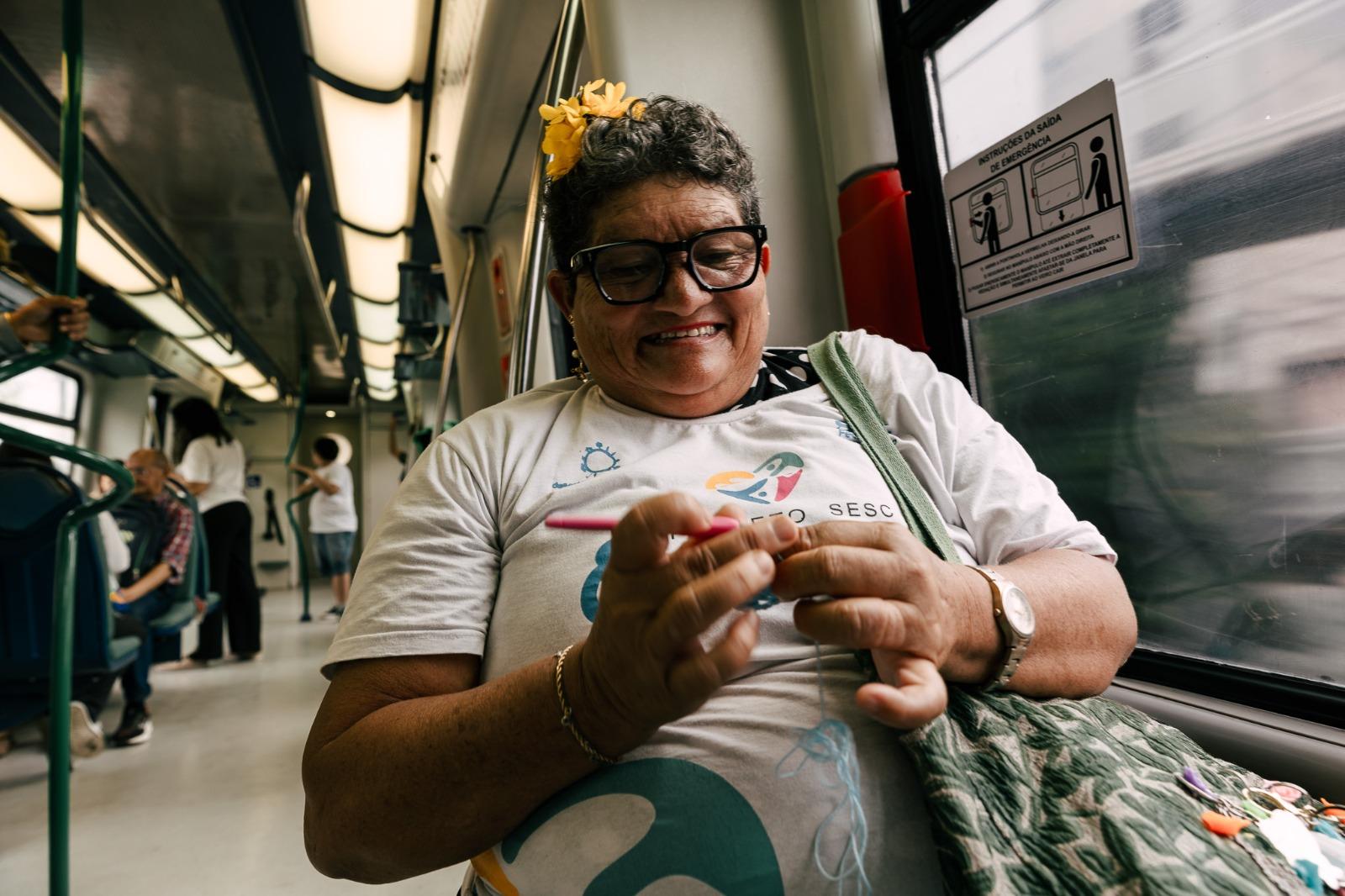 Mulher sorridente sentada dentro de um vagão de metrô, fazendo crochê com linha azul enquanto segura uma agulha rosa, ao lado da janela