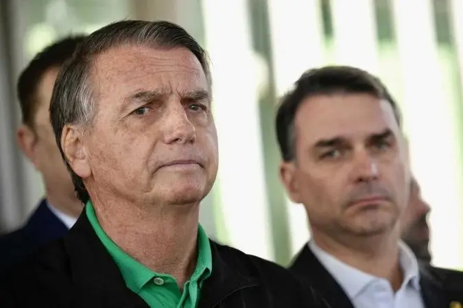 A imagem mostra dois homens em primeiro plano, um à frente do outro. São Jair e Flávio Bolsonaro. O que está mais próximo da câmera tem expressão séria, veste uma camisa verde sob um casaco preto e olha ligeiramente para a esquerda. Atrás dele, um segundo homem, também com semblante sério, usa terno escuro e camisa clara. O fundo é desfocado, com estruturas verticais claras, sugerindo um ambiente externo ou próximo a uma janela ampla.