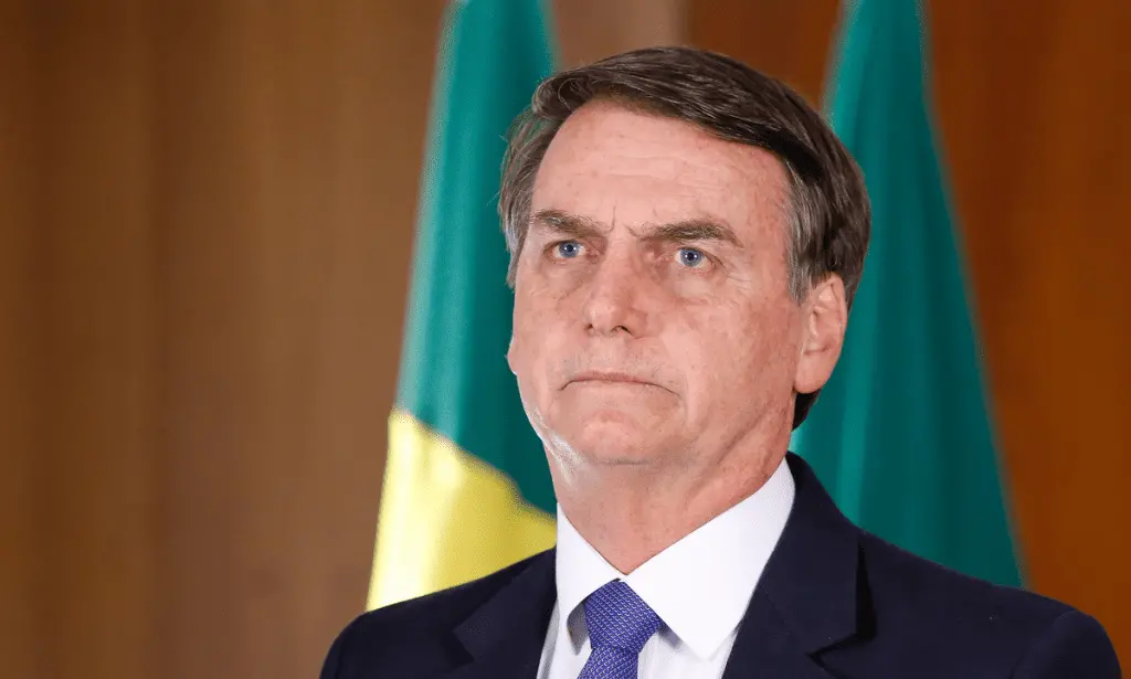 Foto de Jair Bolsonaro, com feição séria, à frente de bandeira do Brasil. Imagem usada para informar que ex-presidente é condenado pelo STF no julgamento da trama golpista