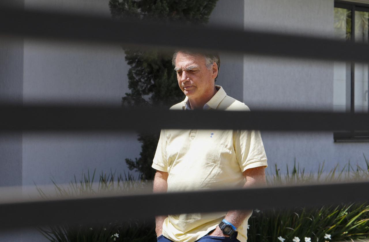 Imagem mostra Jair Bolsonaro cabisbaixo, usando blusa amarela clara, com grades de portão sobre figura dele, enquanto cumpre prisão domiciliar em Brasília