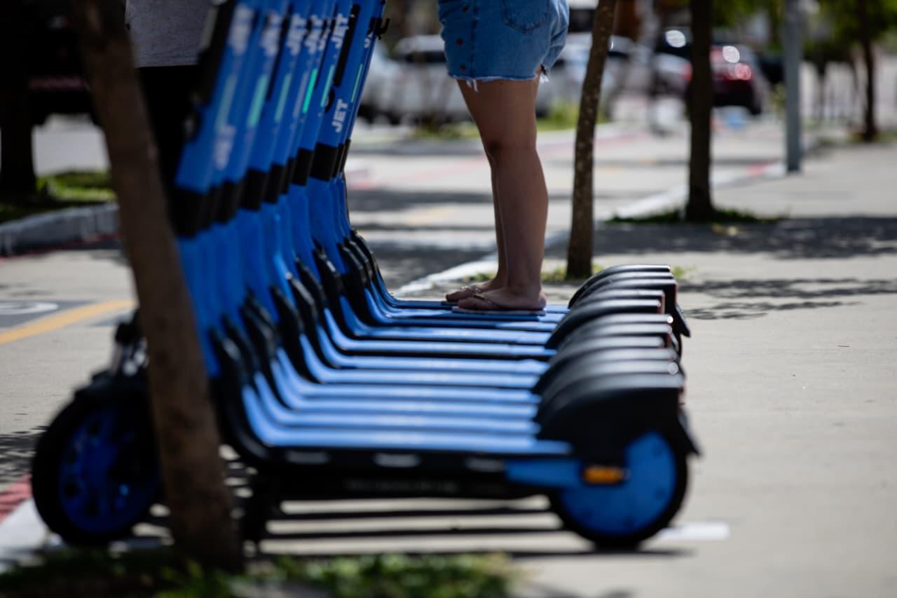 Pernas de pessoa de shorts jeans, equilibrando-se em patinete elétrico azul em calçada, com árvores ao fundo, durante o dia.