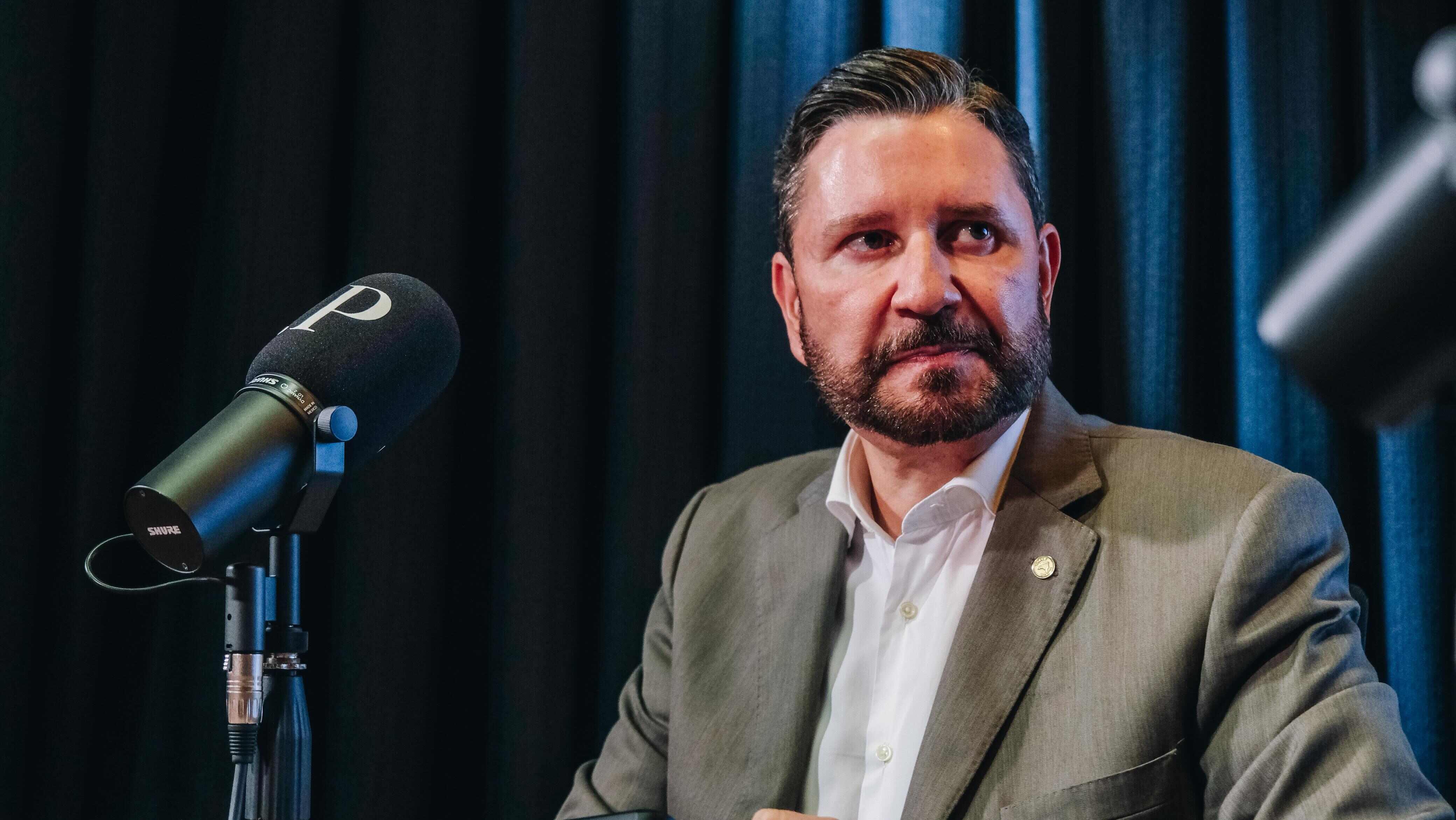 A imagem mostra um homem de barba curta e cabelo penteado para trás, vestindo terno cinza e camisa branca, sentado diante de um microfone preto com capa de espuma onde há a letra “P”. O fundo é composto por cortinas escuras, criando um cenário típico de estúdio de gravação ou podcast.