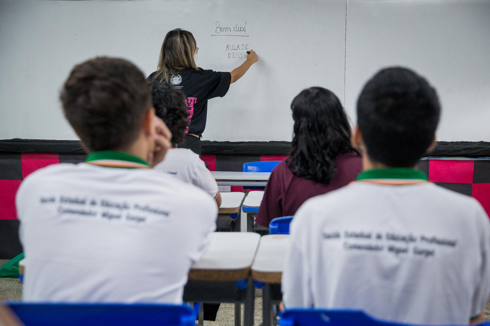 Imagem mostra a visão de trás de uma sala de aula. Uma professora, de costas, escreve em um quadro branco. Alunos estão sentados em carteiras, observando. Dois alunos em primeiro plano usam camisetas brancas de fardamento.