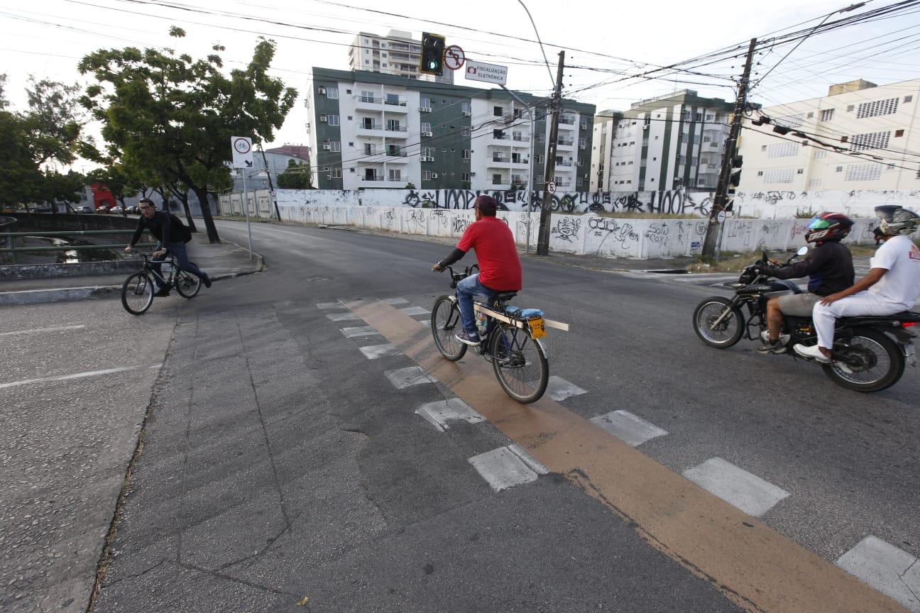 Ciclistas e motociclistas aguardam no cruzamento de uma avenida urbana, próximos a um semáforo, com prédios residenciais ao fundo e muros pichados