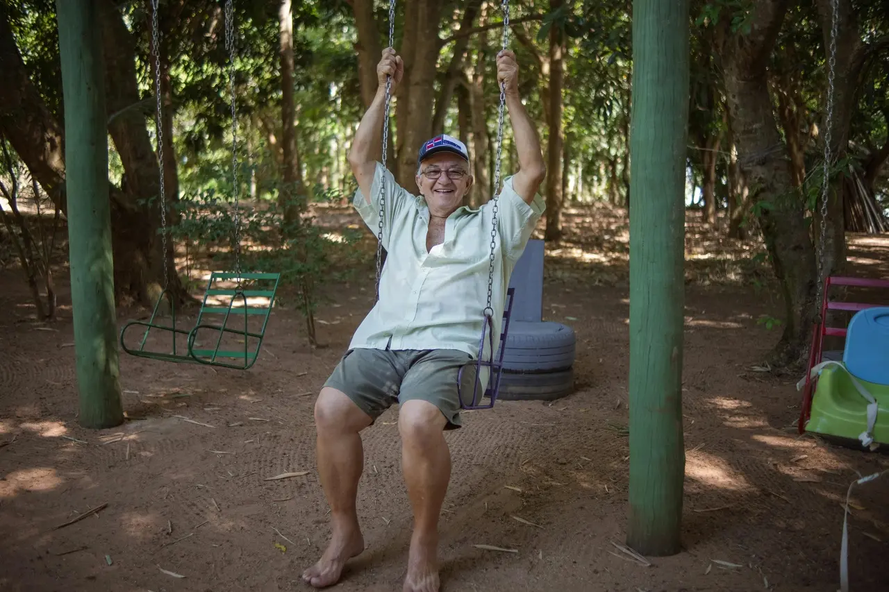 Pessoa idosa sorridente sentada em um balanço, em área arborizada, vestindo camisa clara, bermuda e boné