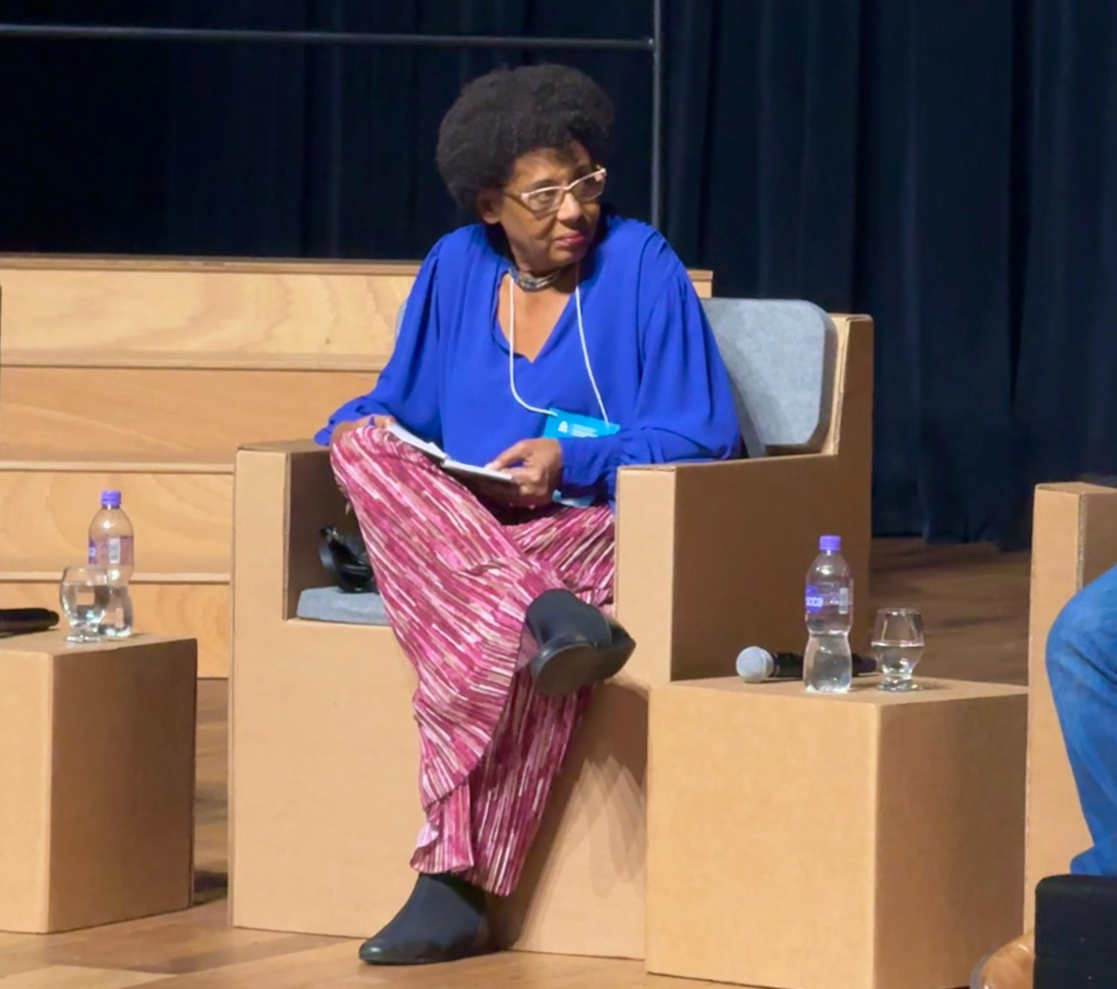 Uma fotografia de uma mulher negra com cabelo curto e crespo e óculos, sentada em uma grande poltrona moderna em um palco. Ela está segurando um caderno e usando uma blusa azul vibrante e calças listradas em rosa e branco. Duas garrafas de água plásticas e um microfone estão em uma pequena caixa ao lado dela.