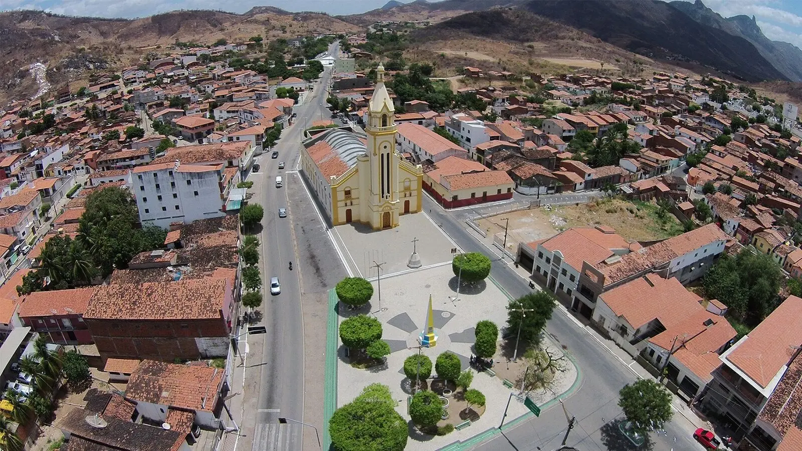 Foto aérea da cidade de Itapajé, no Ceará, com igreja católica amarela ao centro, rodeada de casas, praças e ruas. Ao fundo, alguns montes