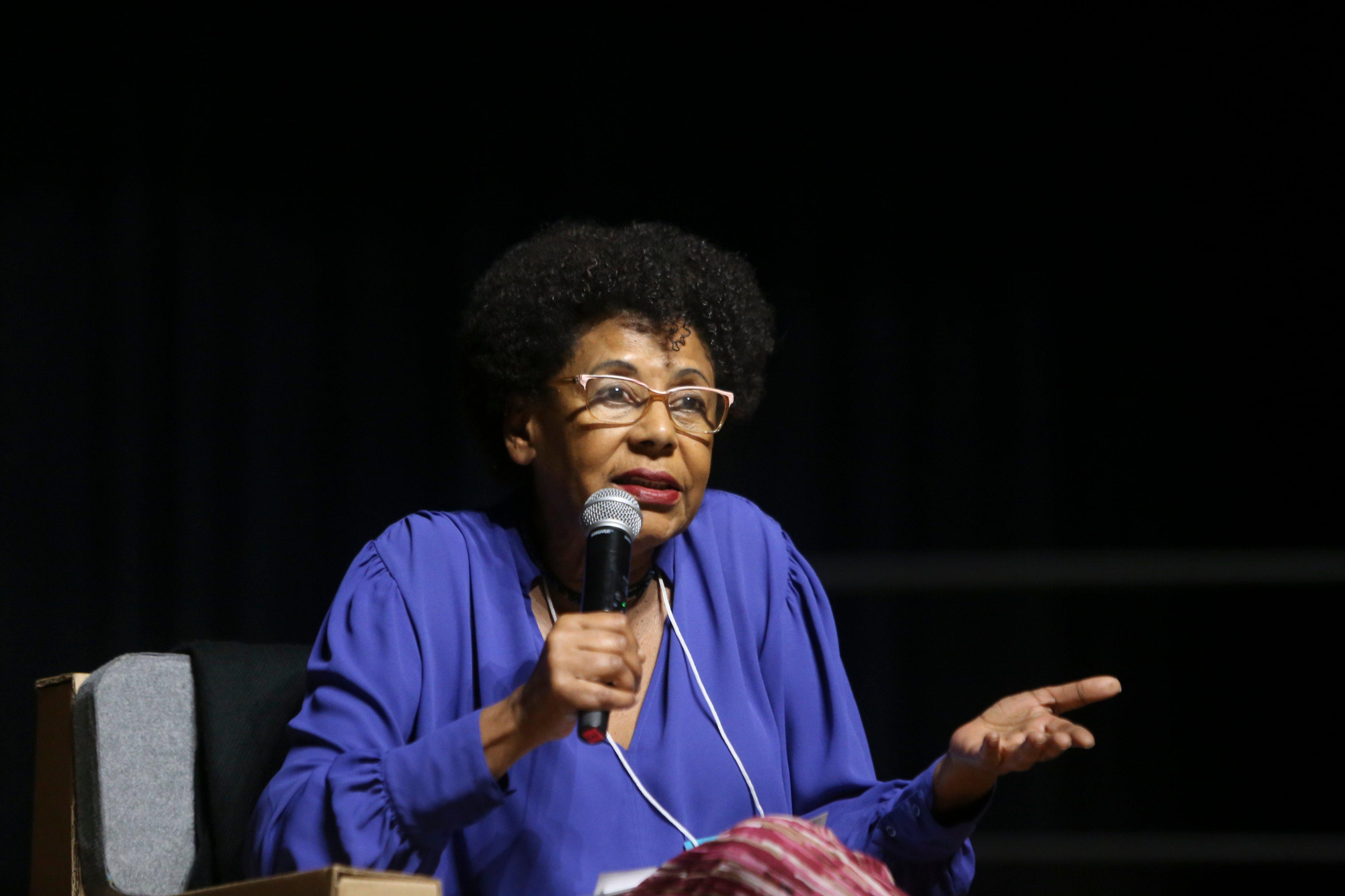 Uma foto em close de Cida Bento, uma mulher negra com cabelos afro curtos, segurando um microfone e gesticulando com a mão esquerda enquanto fala em uma palestra. Ela usa um blusa azul de mangas compridas e óculos de armação clara. O fundo é escuro.