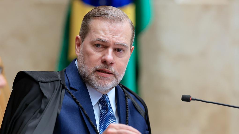 Imagem mostra Toffoli, um homem de meia-idade, com barba grisalha aparada, usando terno azul, camisa clara e gravata azul, sobre o qual veste uma toga preta. Ele gesticula com a mão direita enquanto fala diante de um microfone, em um ambiente formal. Ao fundo, há partes desfocadas de bandeiras nas cores verde, amarela e azul.