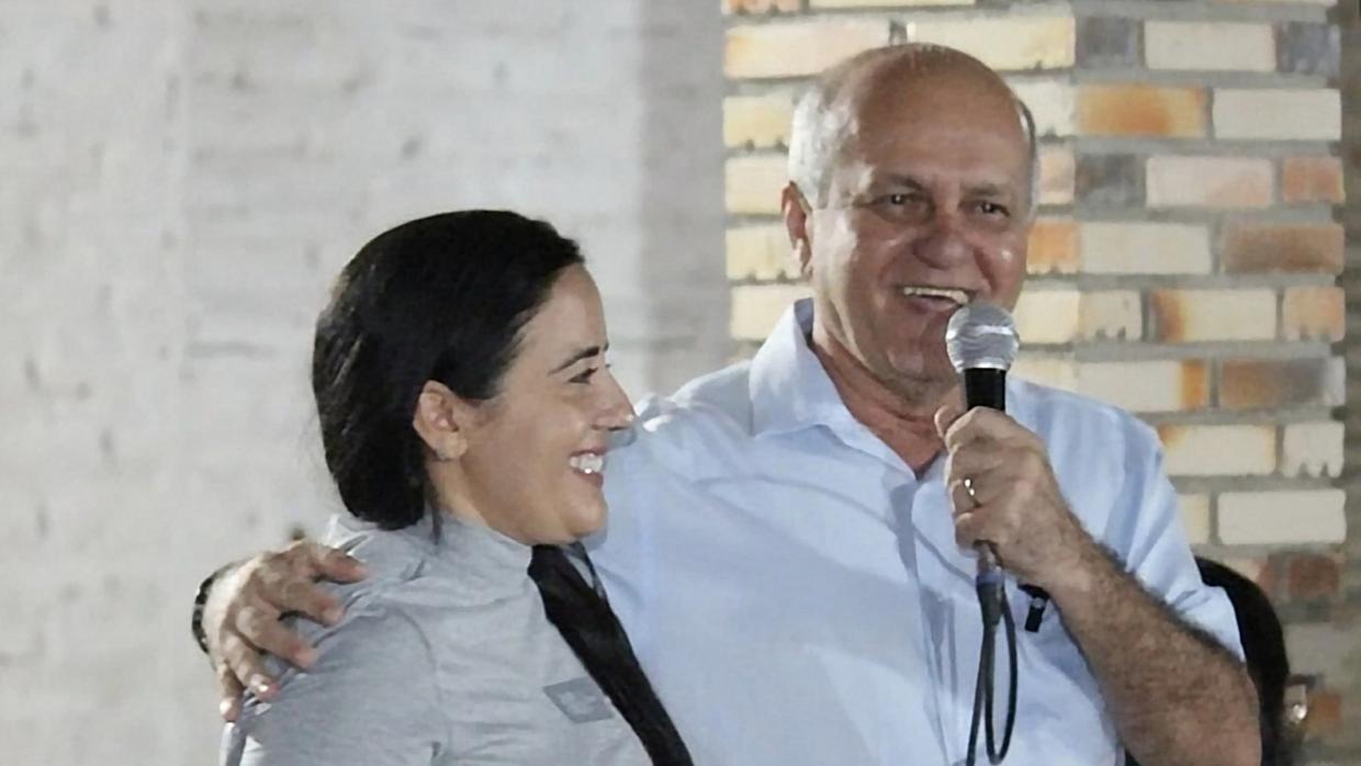 A foto mostra duas pessoas sorrindo em um ambiente interno. Um homem de meia-idade, calvo, veste camisa social azul-clara e segura um microfone enquanto abraça uma mulher de cabelos pretos presos, que usa uma blusa cinza de manga comprida. Ao fundo, há uma parede de tijolos aparentes em tons claros e um trecho de parede branca.