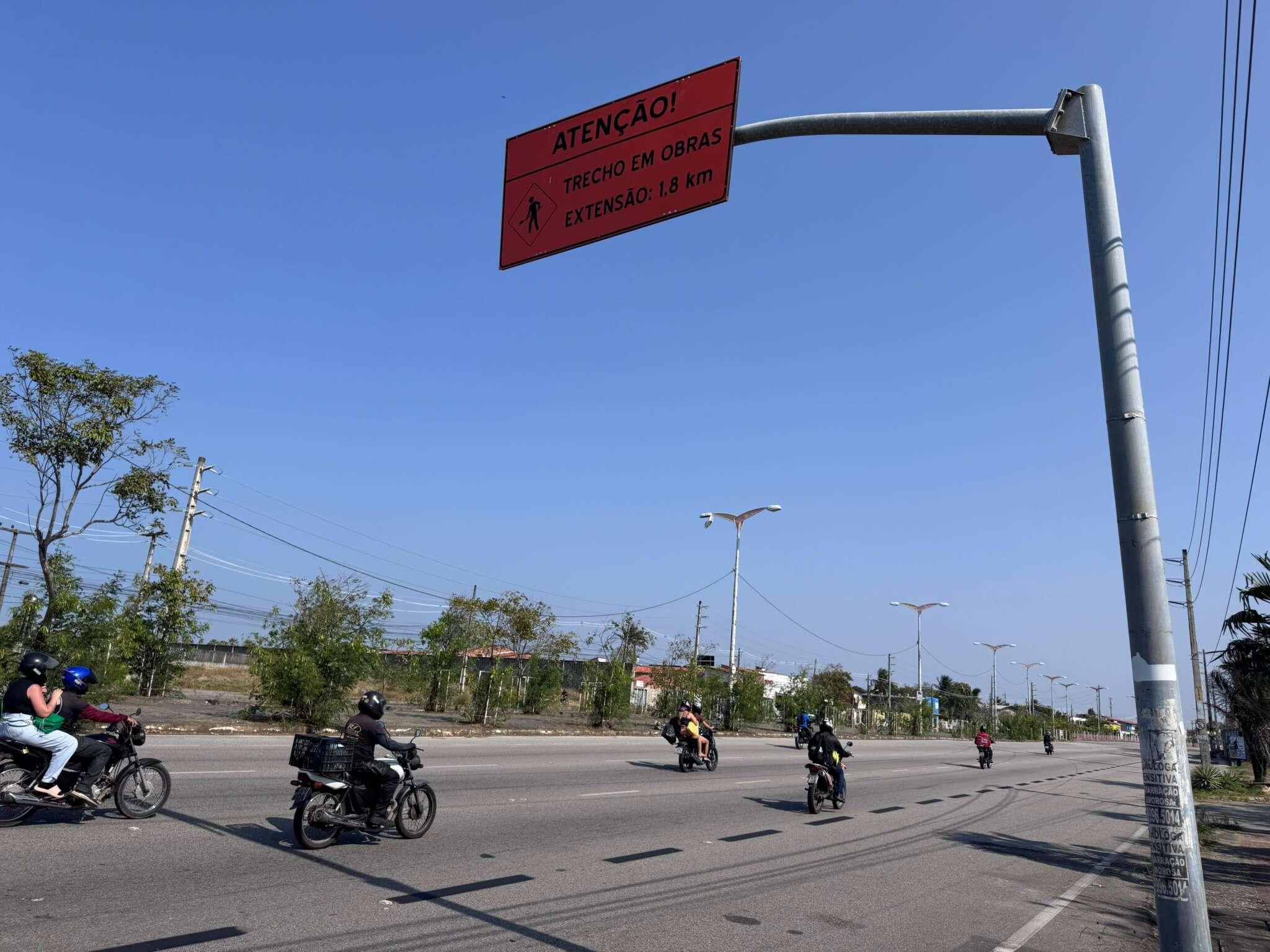 Aviso de obra em avenida, indicando trecho em obras de 1,8 km, com motociclistas passando pelo local, sinalizando atenção e cuidado ao dirigir.