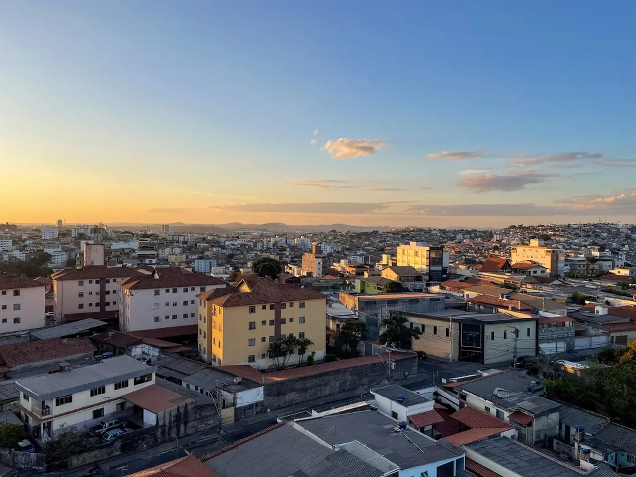 Vista panorâmica da cidade brasileira de Contagem ao entardecer, com prédios variados e céu com nuvens e tonalidades quentes do pôr do sol.