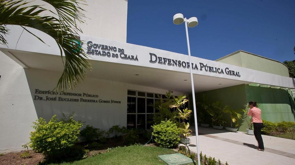 Fachada da Defensoria Pública Geral do Estado do Ceará, com o letreiro “Governo do Estado do Ceará – Defensoria Pública Geral” e “Edifício Defensor Público Dr. José Euclides Ferreira Gomes Júnior”. O prédio tem paredes brancas e verdes, jardim com plantas ornamentais e uma pessoa de camisa laranja caminhando na calçada em direção à entrada.