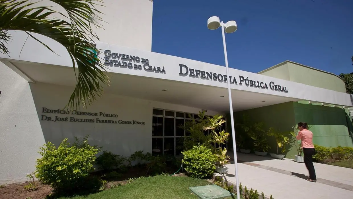 Fachada da Defensoria Pública Geral do Estado do Ceará, com o letreiro “Governo do Estado do Ceará – Defensoria Pública Geral” e “Edifício Defensor Público Dr. José Euclides Ferreira Gomes Júnior”. O prédio tem paredes brancas e verdes, jardim com plantas ornamentais e uma pessoa de camisa laranja caminhando na calçada em direção à entrada.