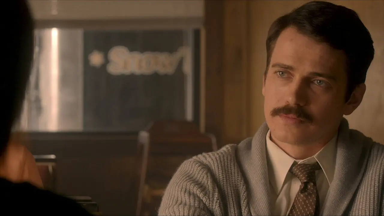 Imagem de um homem com expressão pensativa, trajando camisa social, gravata e suéter, em ambiente com iluminação suave e decoração de bar ou restaurante.