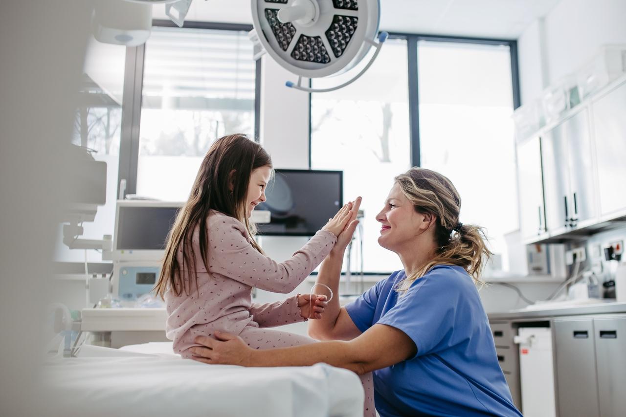 Imagem de quarto de hospital. Uma menina sentada na cama, com pijama rosa e cabelos longos, de frente a uma médica de roupa azul e cabelos claros preso. Elas sorriem e tocam uma a mão da outra