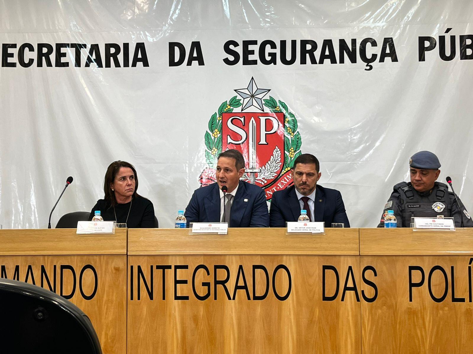 Coletiva de imprensa da Secretaria da Segurança Pública de São Paulo. Quatro autoridades estão sentadas à mesa: uma mulher, dois homens de terno e um policial militar fardado. Ao fundo, painel com o brasão do estado de São Paulo e a inscrição 'Secretaria da Segurança Pública'.