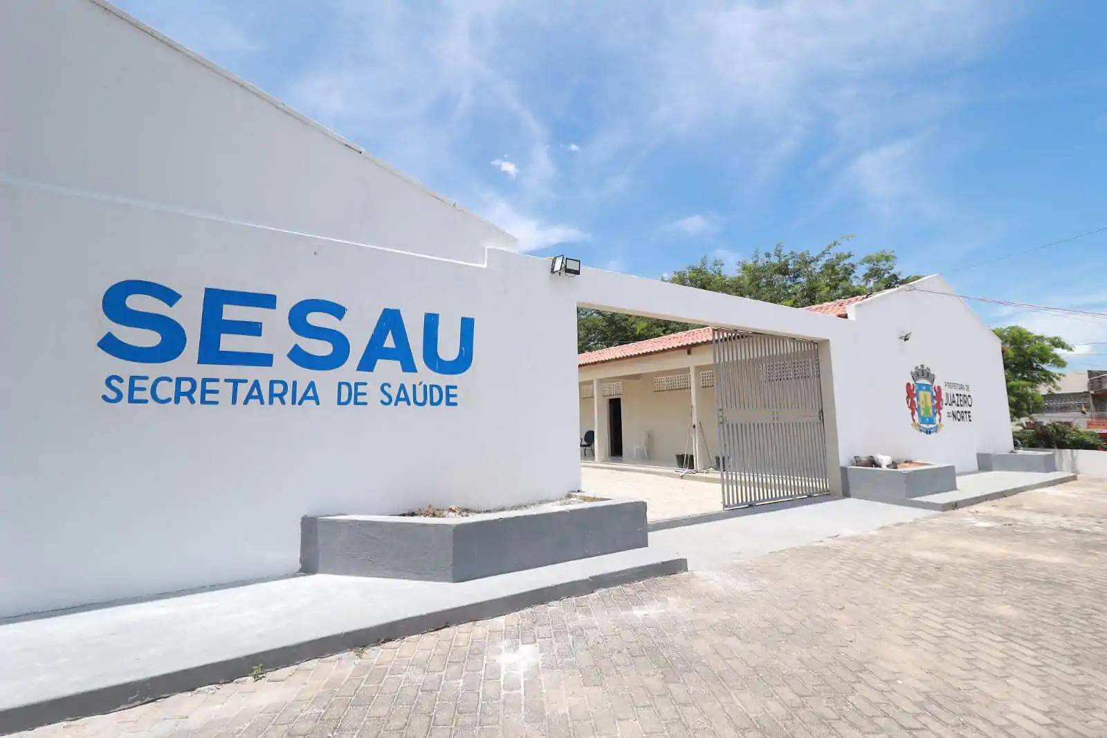achada da Secretaria de Saúde de Juazeiro do Norte (SESAU), com paredes brancas, letreiro azul e o brasão da prefeitura ao lado do portão de entrada