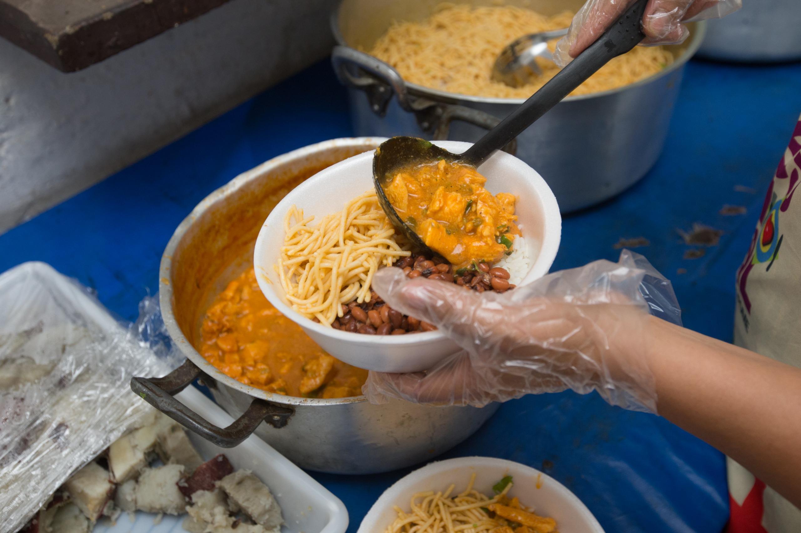 Detalhe de uma mão com luva de plástico servindo macarrão, feijão e um ensopado de um pote grande para um pequeno prato branco.