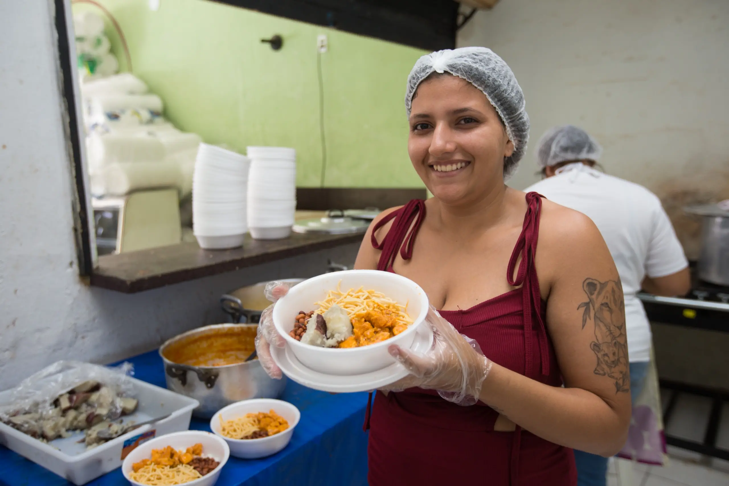 Uma mulher sorri para a câmera enquanto segura um prato branco cheio de comida, mostrando uma refeição já servida. Ela usa um vestido vinho, rede de cabelo e luvas.