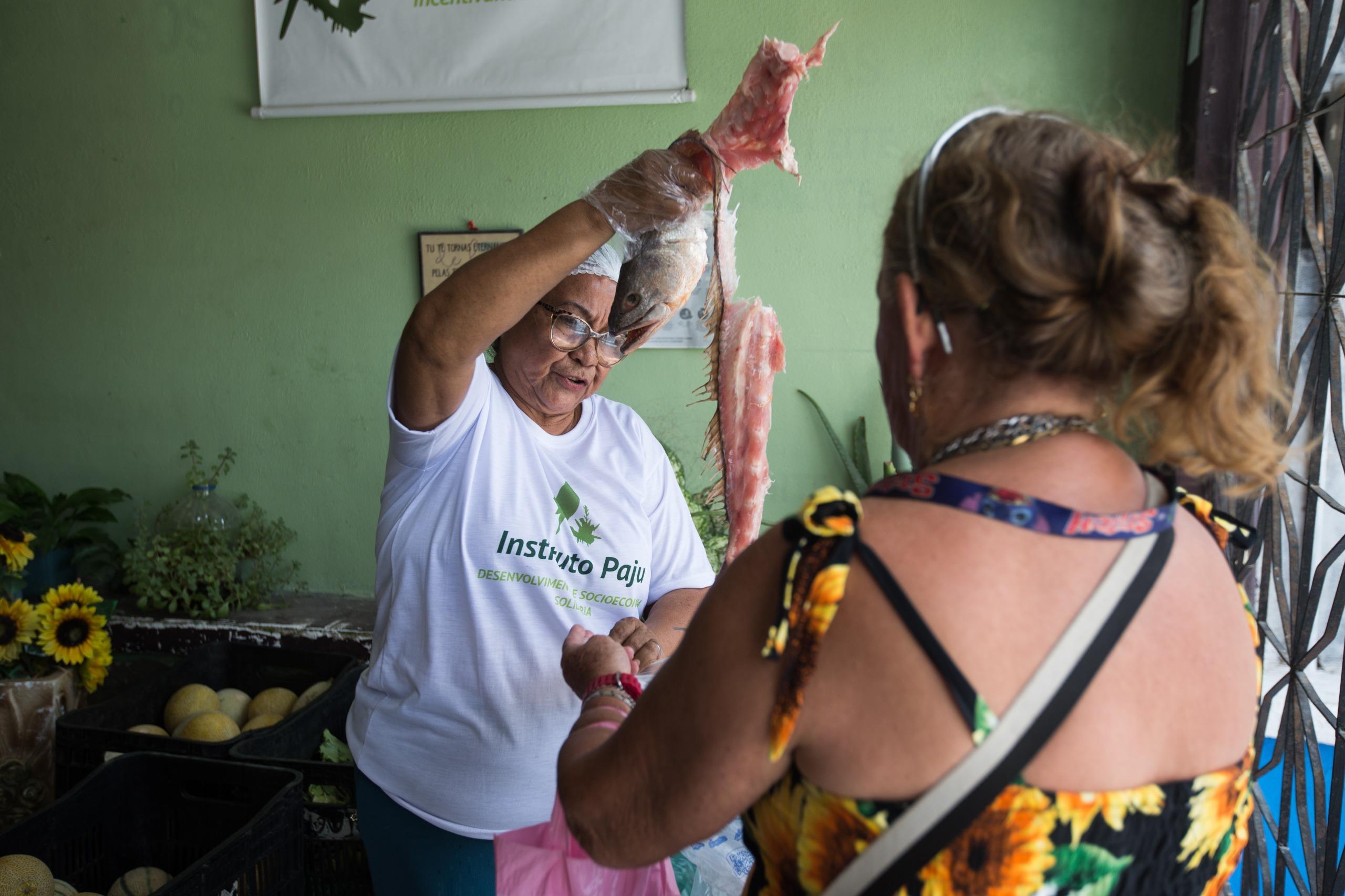 Uma mulher baixa, de touca e blusa branca, coloca uma carcaça de peixe dentro da sacola segurada por outra mulher, de costas.