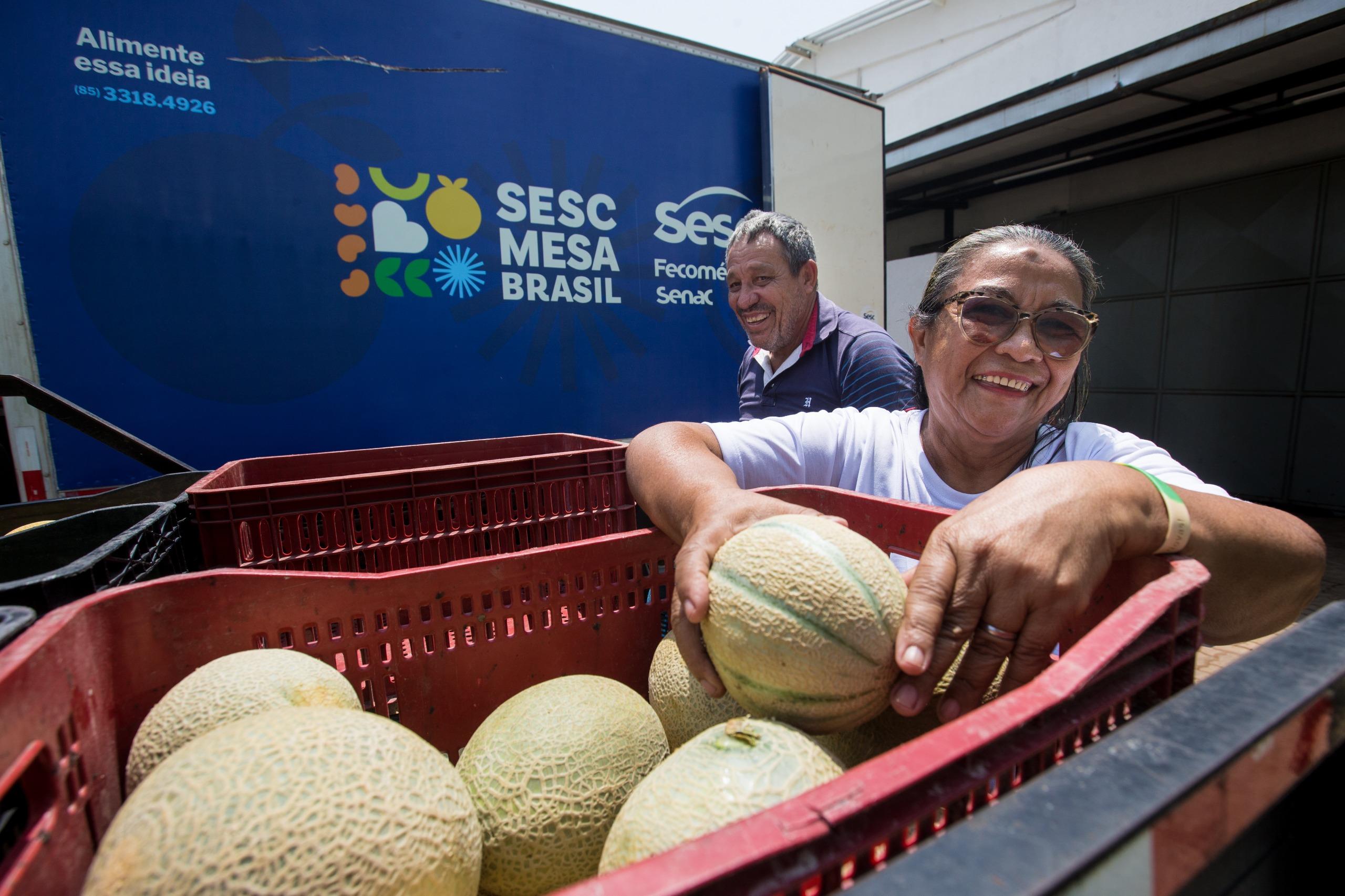 Imagem mostra mulher de óculos retirando um melão de um engradado vermelho. Ao fundo, um homem sorri, próximo a um caminhão azul do Sesc Mesa Brasil