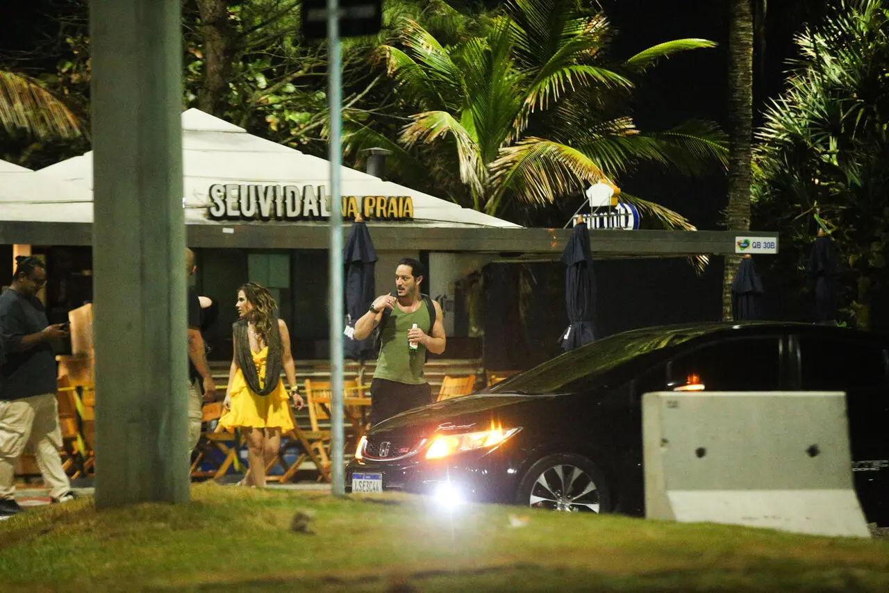 Wanessa, de vestido amarelo, e Allan, de blusa verde e calça preta, indo embora no mesmo veículo. Vegetação tropical ao fundo, perto de um carro preto e uma estrutura de bar.