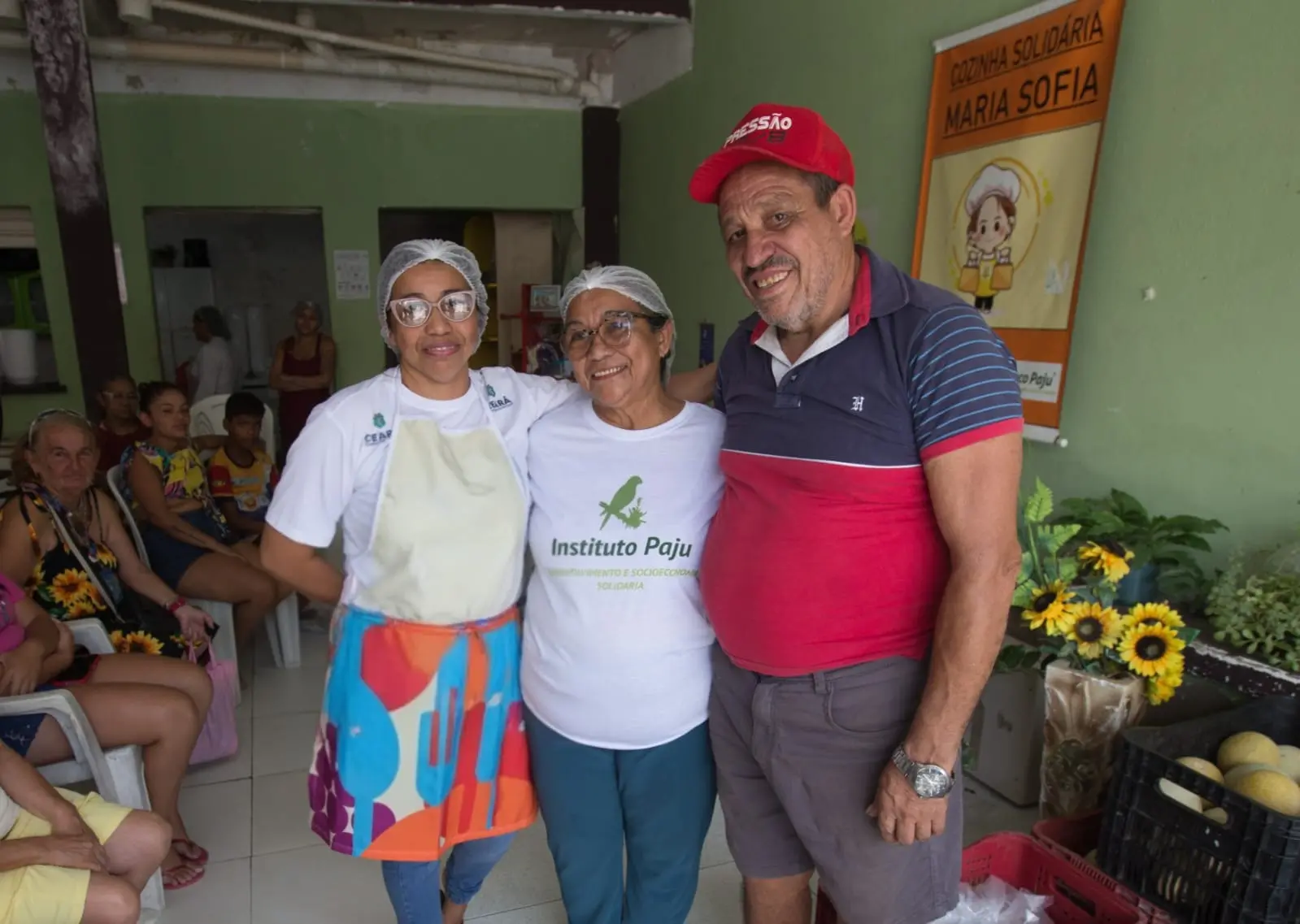 Duas mulheres e um homem posam para a foto. A primeira está de avental, a segunda de camisa branca, e o homem veste uma bermuda jeans, camisa azul com vermelho e boné.