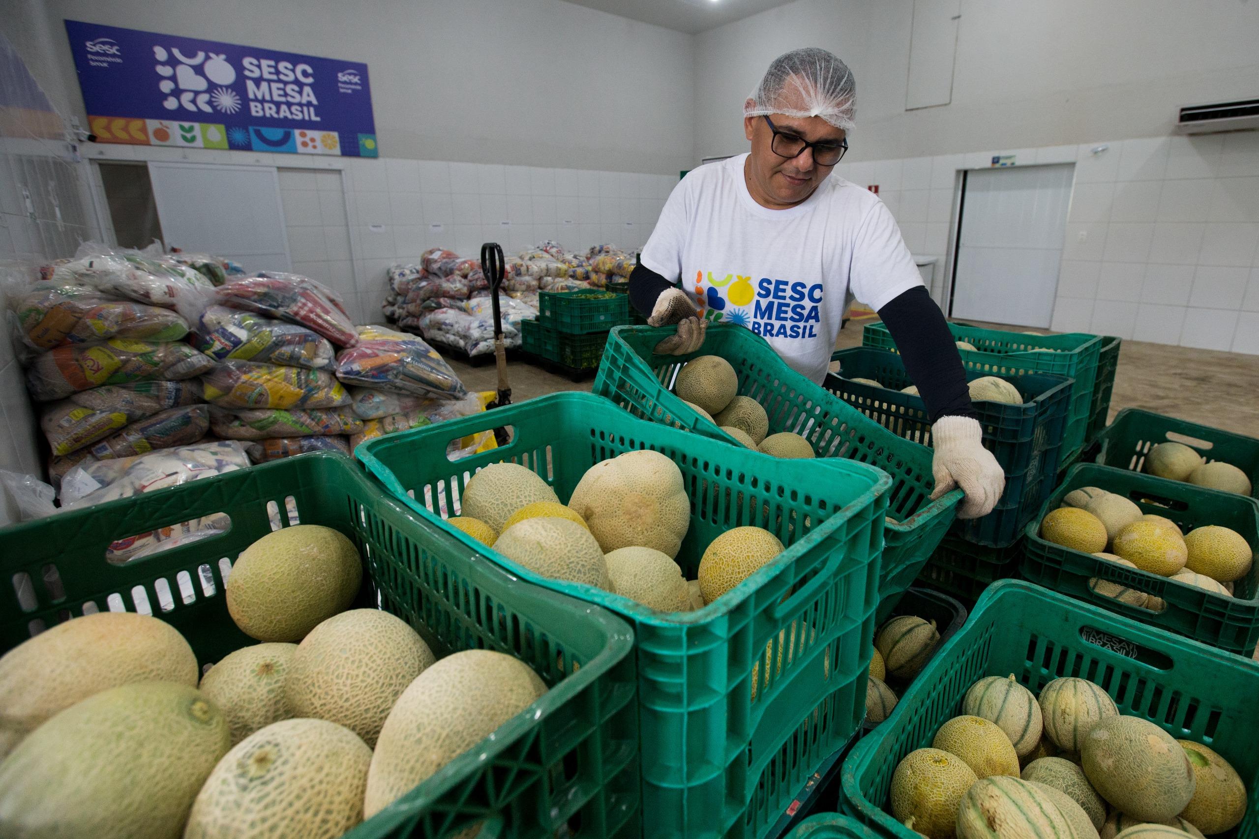 Um homem de touca e luvas, usando uma camiseta com a inscrição Sesc Mesa Brasil, organiza caixas plásticas verdes cheias de melões. Ao fundo, sacos de alimentos estão empilhados.