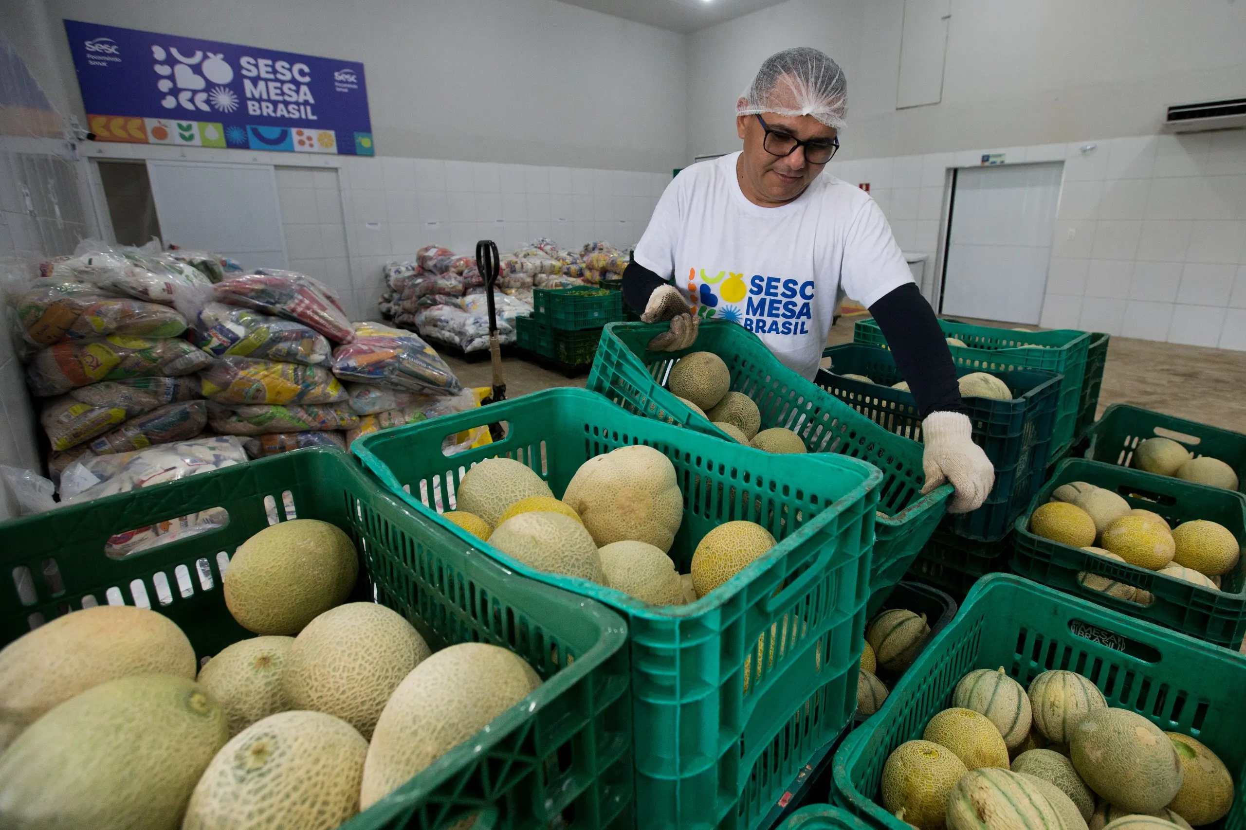 Um homem de touca e luvas, usando uma camiseta com a inscrição Sesc Mesa Brasil, organiza caixas plásticas verdes cheias de melões. Ao fundo, sacos de alimentos estão empilhados.