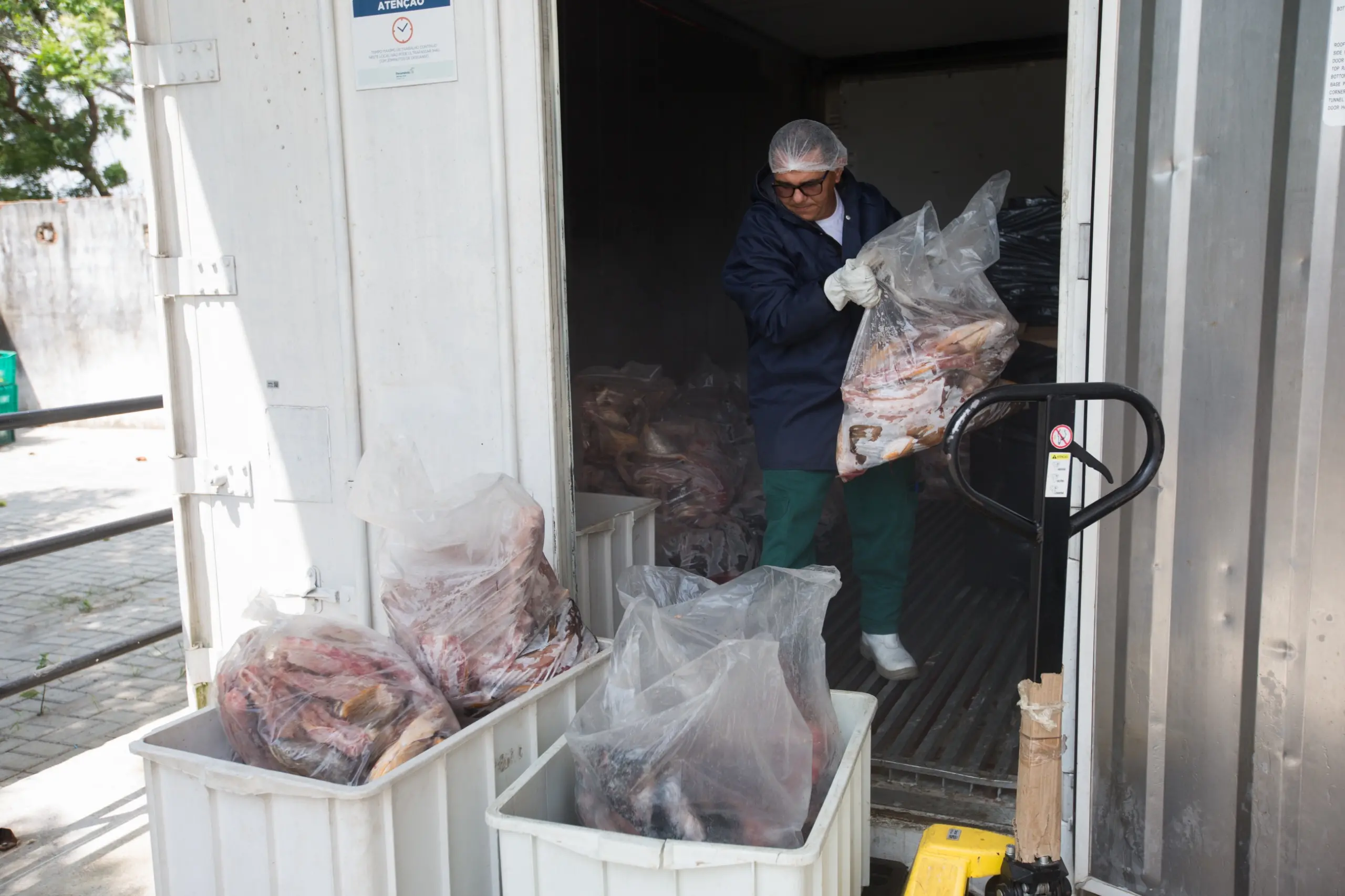 Um homem vestido de touca, luvas e avental está dentro de uma câmara fria carregando sacos de peixe congelado
