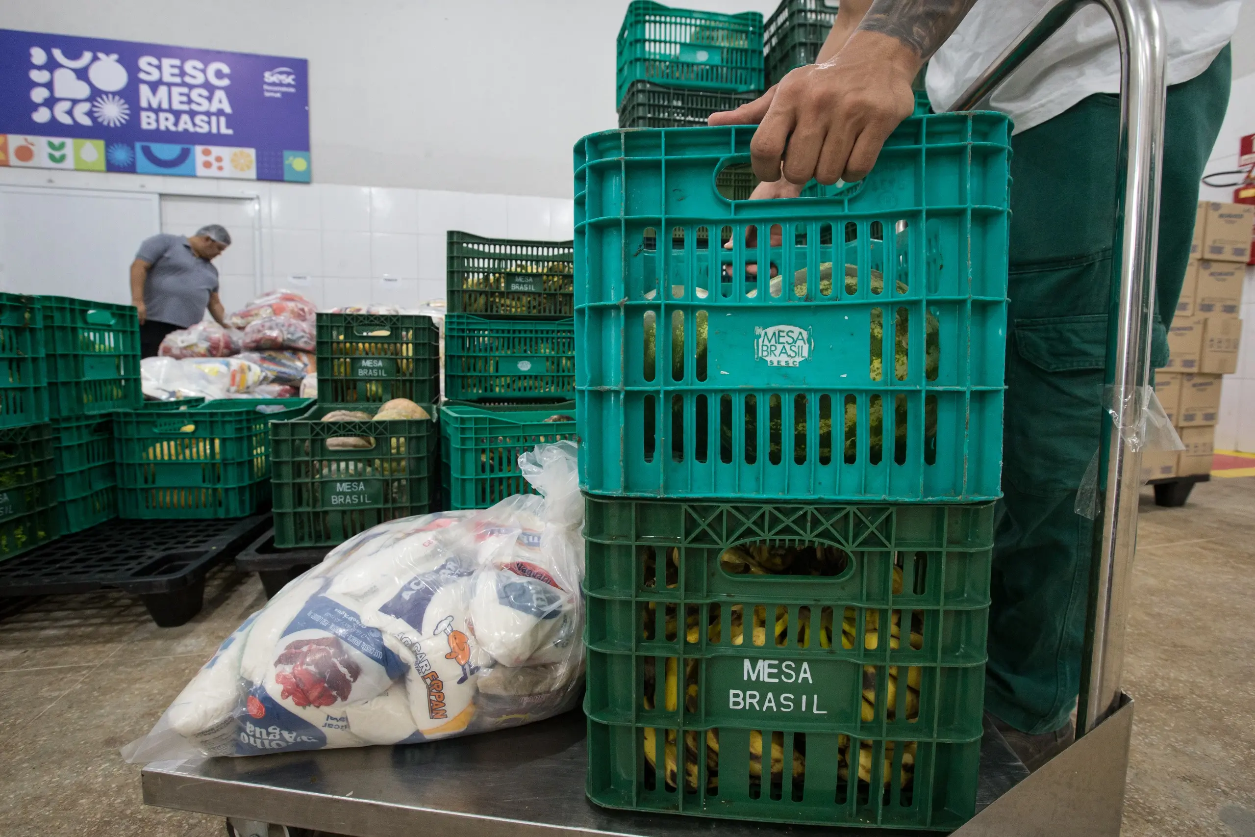 Imagem mostra uma mão empilhando engradados verdes, cheios de frutas. Ao lado, uma sacola grande com vários quilos de alimentos em um galpão.