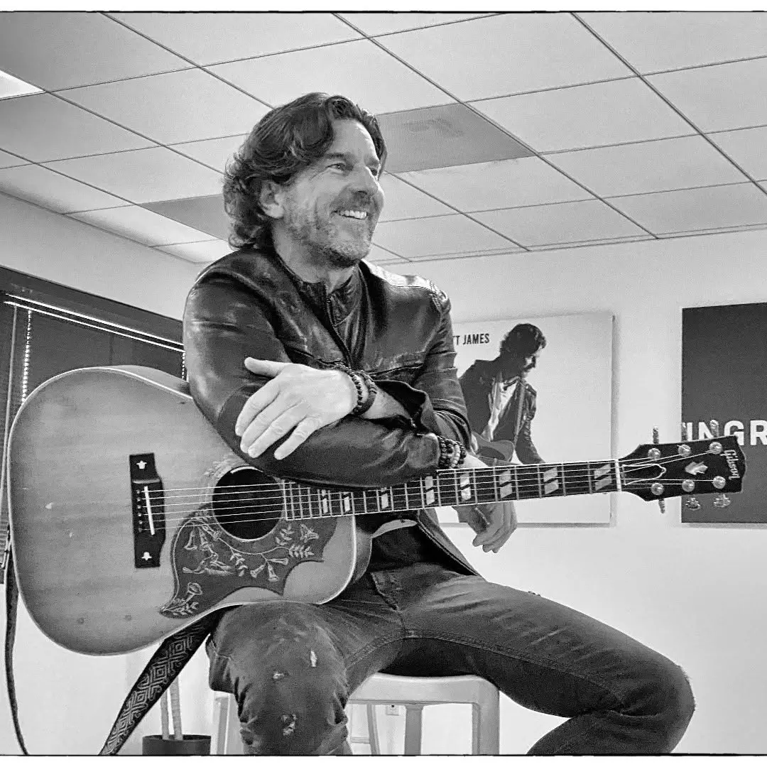 Imagem, em escala de cinza, mostra o compositor Brett James, ganhador do Grammy, sentado em um banco enquanto se apoia sobre um violão no seu colo e sorrir.