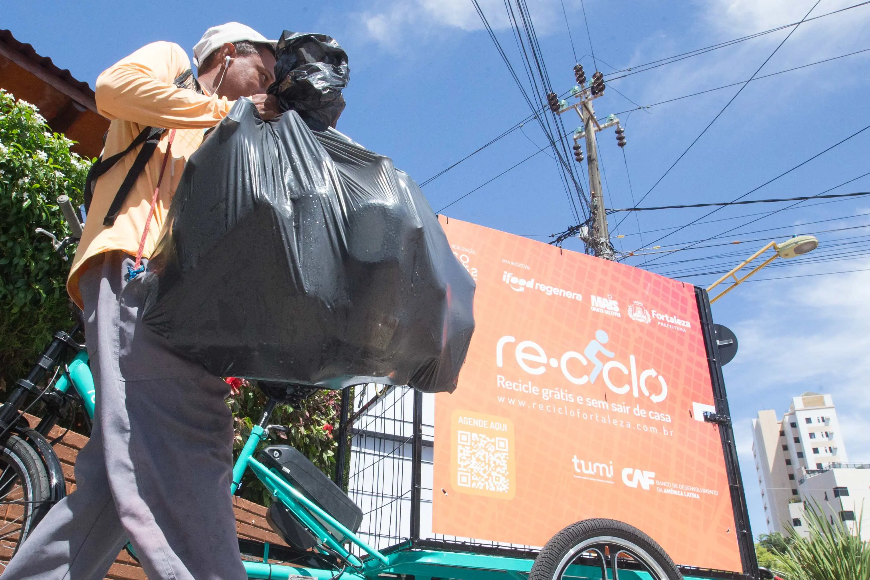 Um catador de materiais recicláveis, vestindo camisa laranja e boné, coloca um grande saco de lixo preto em um triciclo de carga. Na lateral do triciclo, um painel laranja divulga o projeto 