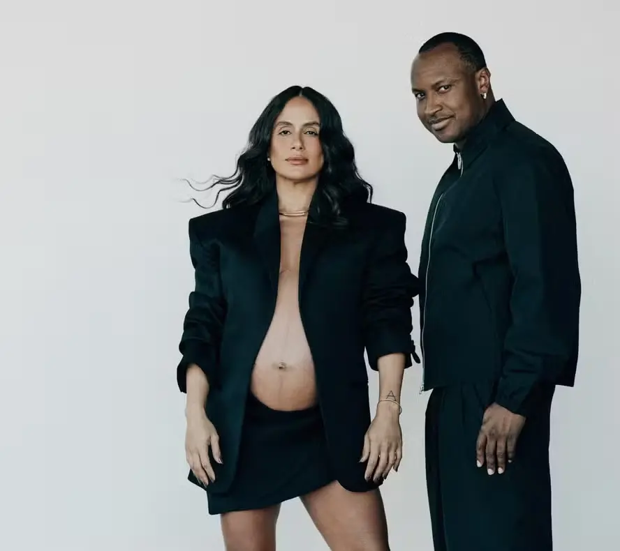 A mulher está grávida e exibe sua barriga descoberta de forma elegante e confiante. Ela veste um blazer preto estruturado, sem blusa por baixo, e uma saia curta preta. Ao lado dela, o homem veste. Ele olha para a câmera com um leve sorriso. Sua expressão é séria e firme, transmitindo força e serenidade.