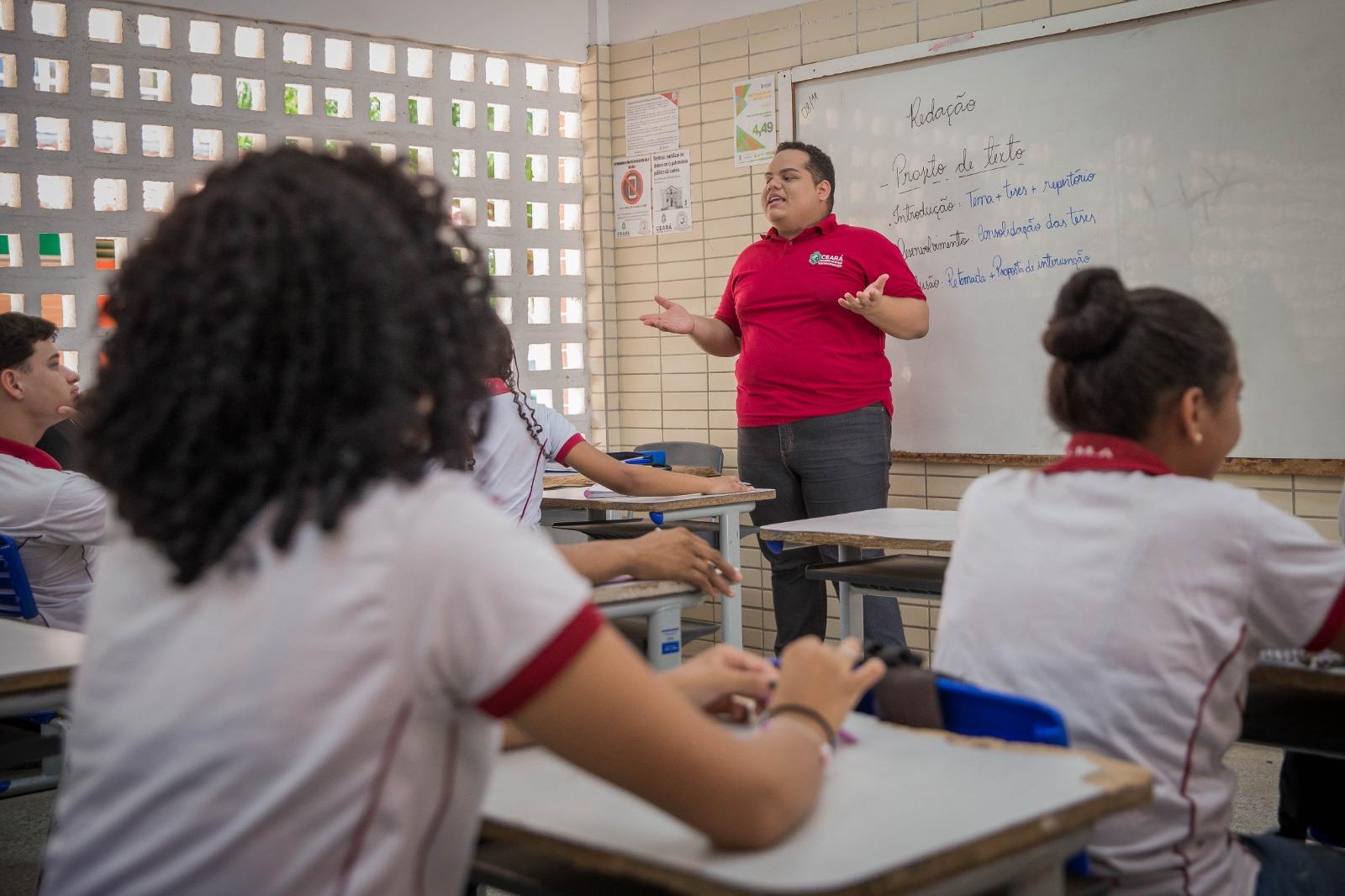 Imagem de uma sala de aula com alunas sentadas de frente para o quadro e um professor posicionado em frente ao quadro usando blusa vermelha