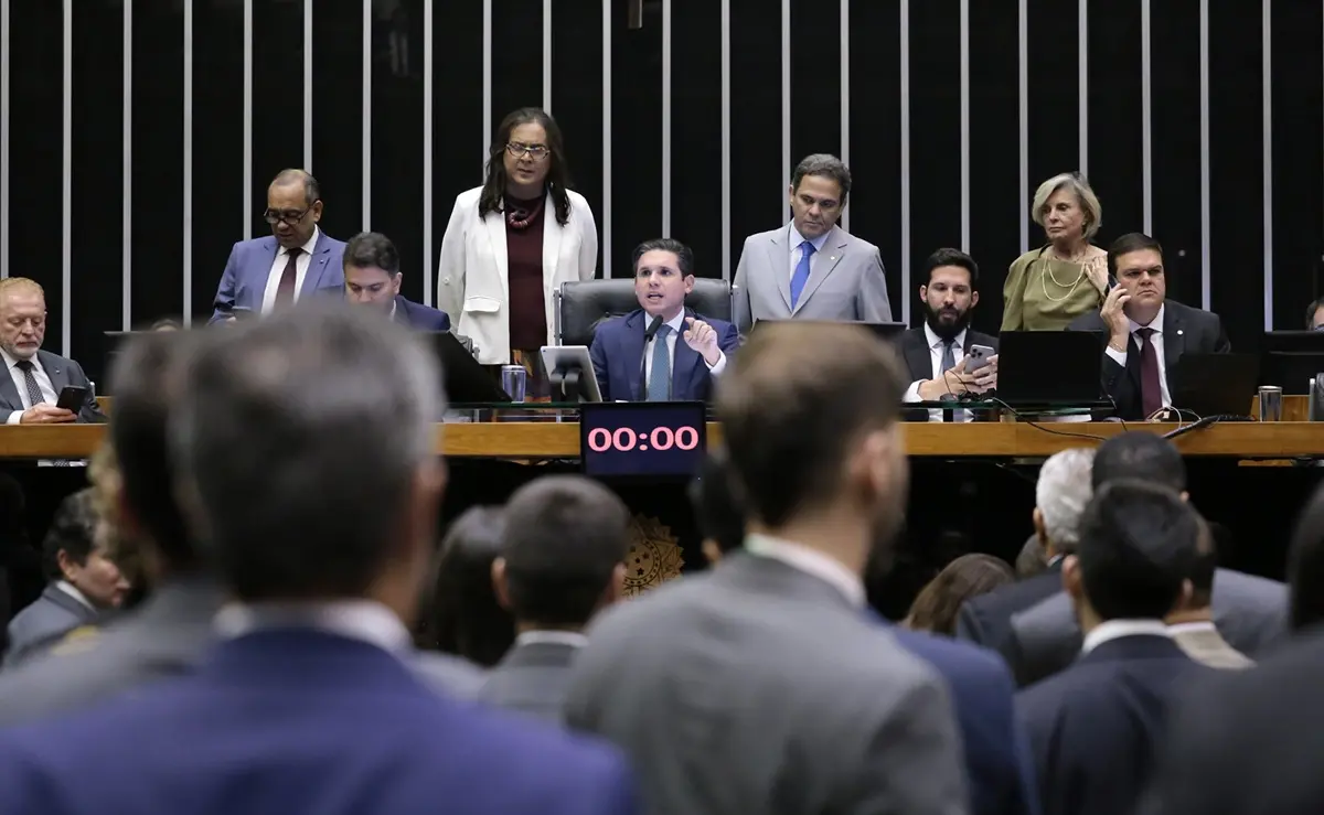 Parlamentares reunidos no plenário da Câmara dos Deputados, com um grupo de deputados à mesa diretora. Um deputado ao centro, sentado, fala ao microfone enquanto outros parlamentares observam, usam celular ou conversam. No painel eletrônico à frente, o cronômetro exibe “00:00”. Na frente da imagem, aparecem de costas alguns deputados assistindo à sessão.