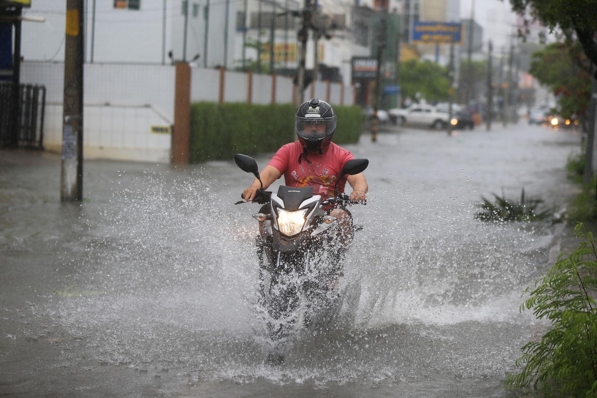 Homem de moto atravessa rua alagada pela chuva em Fortaleza, com água cobrindo parte da pista.