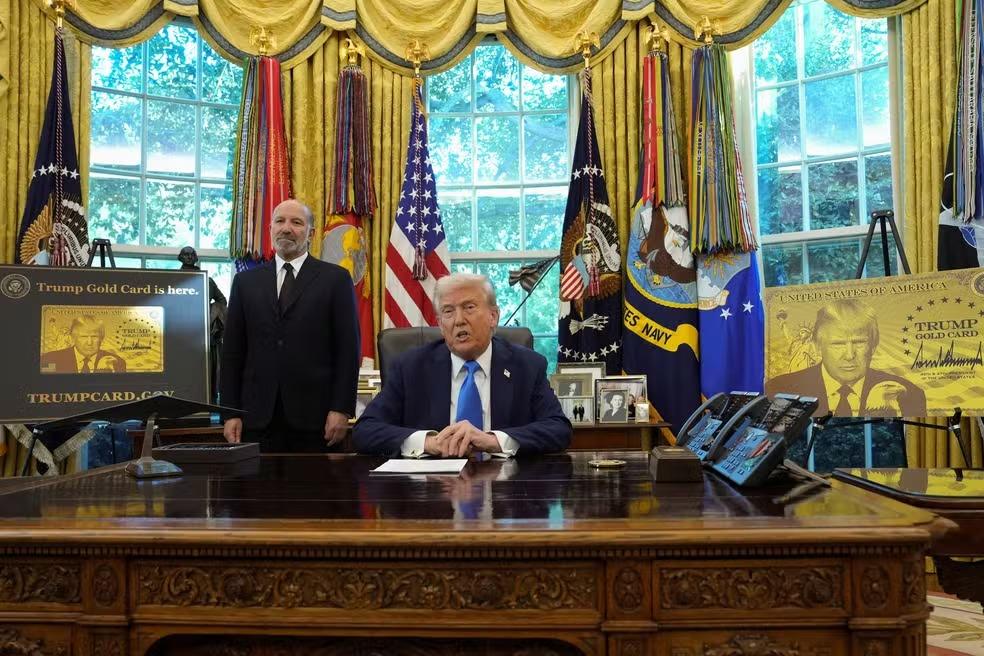 Donald Trump aparece sentado à mesa no Salão Oval da Casa Branca, ao lado de um homem em pé.