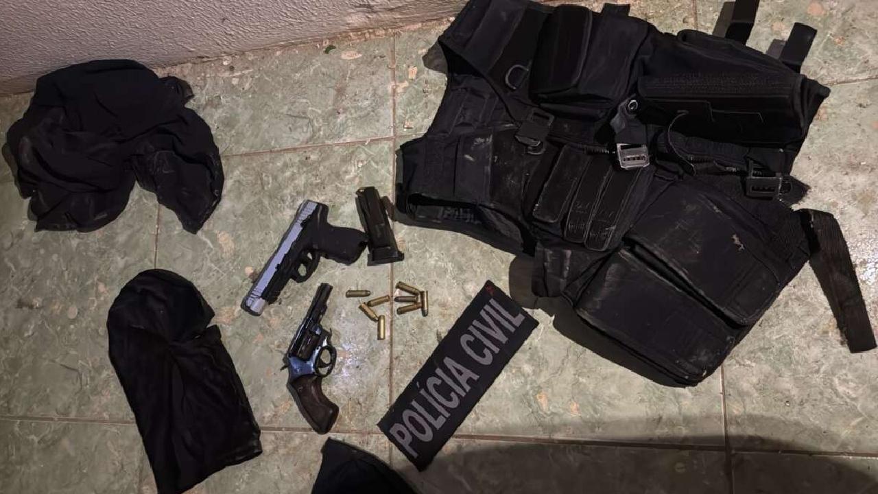 Duas armas de fogo, munições, carregador, colete tático preto e balaclavas pretas estão dispostos no chão de cerâmica, ao lado de um brasão escrito 