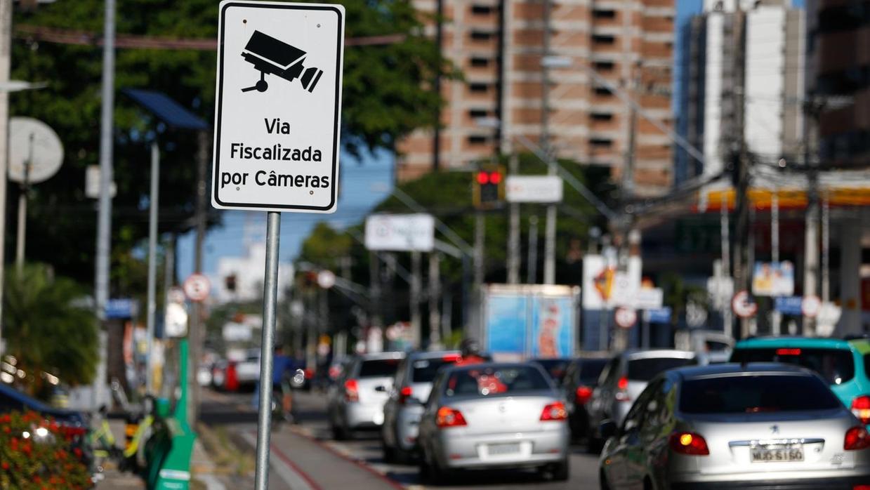 Foto que contém veículos parados no sinal ao fundo, enquanto na frente o destaque é para placa com a frase 'via fiscalizada por câmeras'