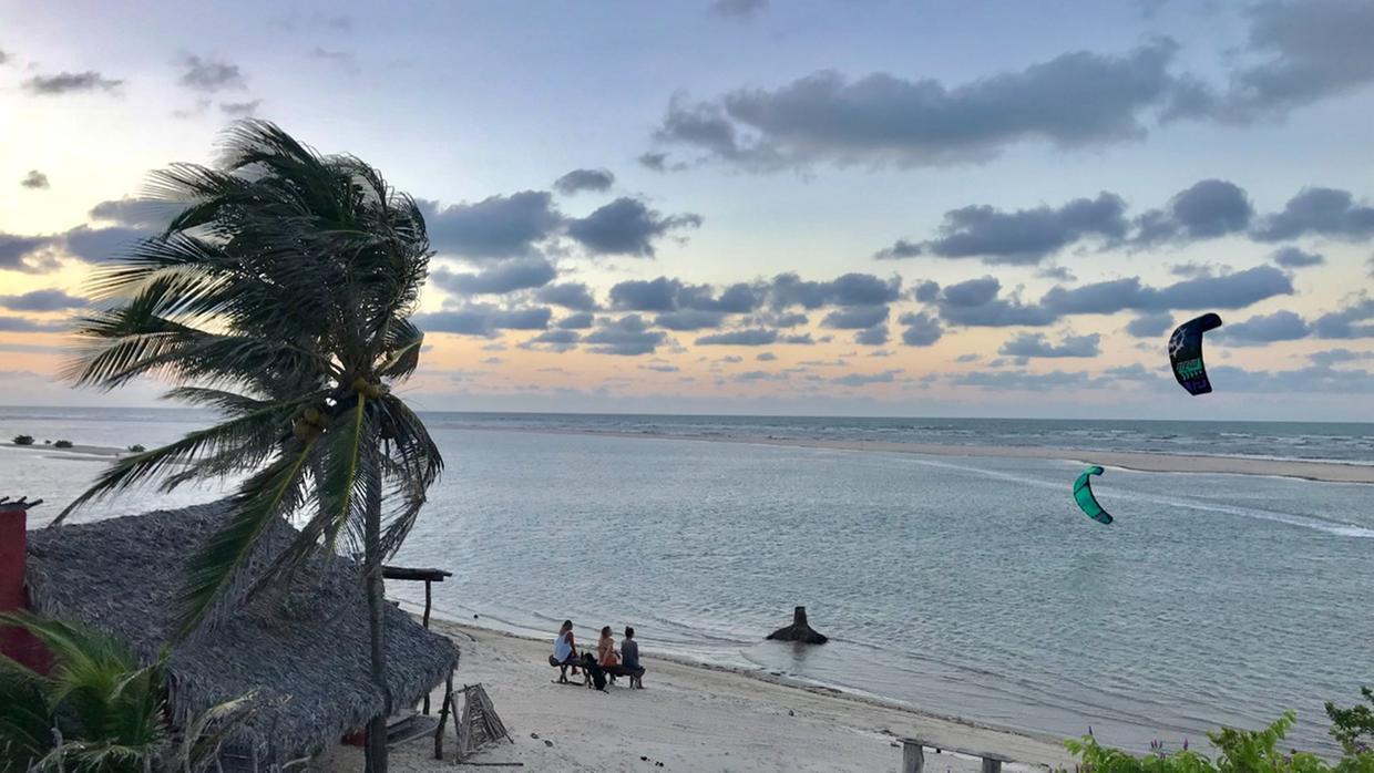 Imagem mostra praia de Tatajuba, em Camocim, no final da tarde, com kitesurfistas praticando esporte e pessoas sentadas à beira do mar