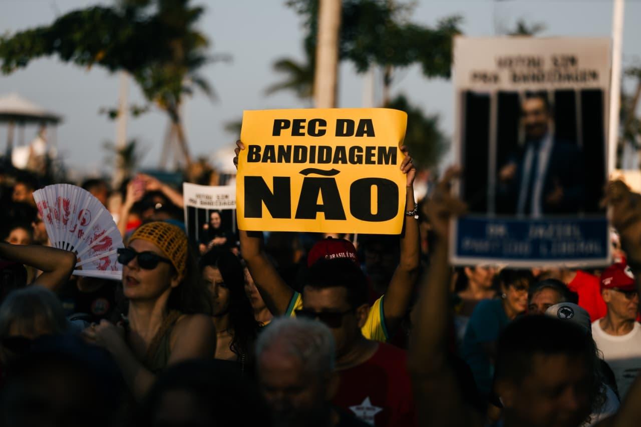 Manifestação popular com muitas pessoas reunidas, algumas segurando cartazes e bandeiras do Brasil. No centro da imagem, um cartaz em destaque diz: “LUGAR DE GOLPISTA É NA CADEIA! PAPUDA”. Outras pessoas ao redor erguem os punhos ou tiram fotos. Ao fundo, vê-se a estátua de Iracema Guardiã