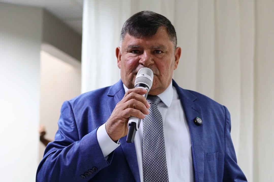 A imagem mostra Braguinha, um homem de meia-idade, com cabelo curto escuro e grisalho, vestindo terno azul, camisa branca e gravata estampada em preto e branco. Ele segura um microfone prateado próximo à boca, como se estivesse discursando. Ao fundo, há uma cortina branca e parte de uma parede clara, sugerindo que a cena ocorre em um ambiente interno, possivelmente durante um evento ou reunião formal.