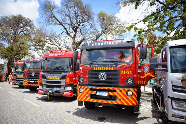 Fileira de carros do Corpo de Bombeiros em estacionamento durante o dia, com árvores ao fundo.