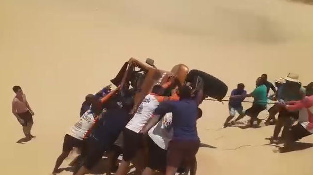 Grupo de pessoas se unindo para desvirar um carro off-road tombado em uma duna de areia