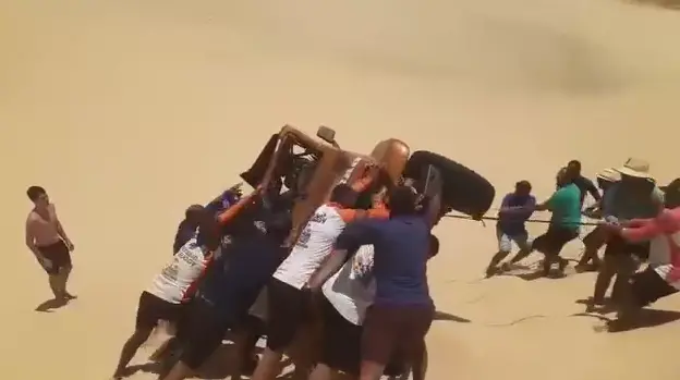 Grupo de pessoas se unindo para desvirar um carro off-road tombado em uma duna de areia