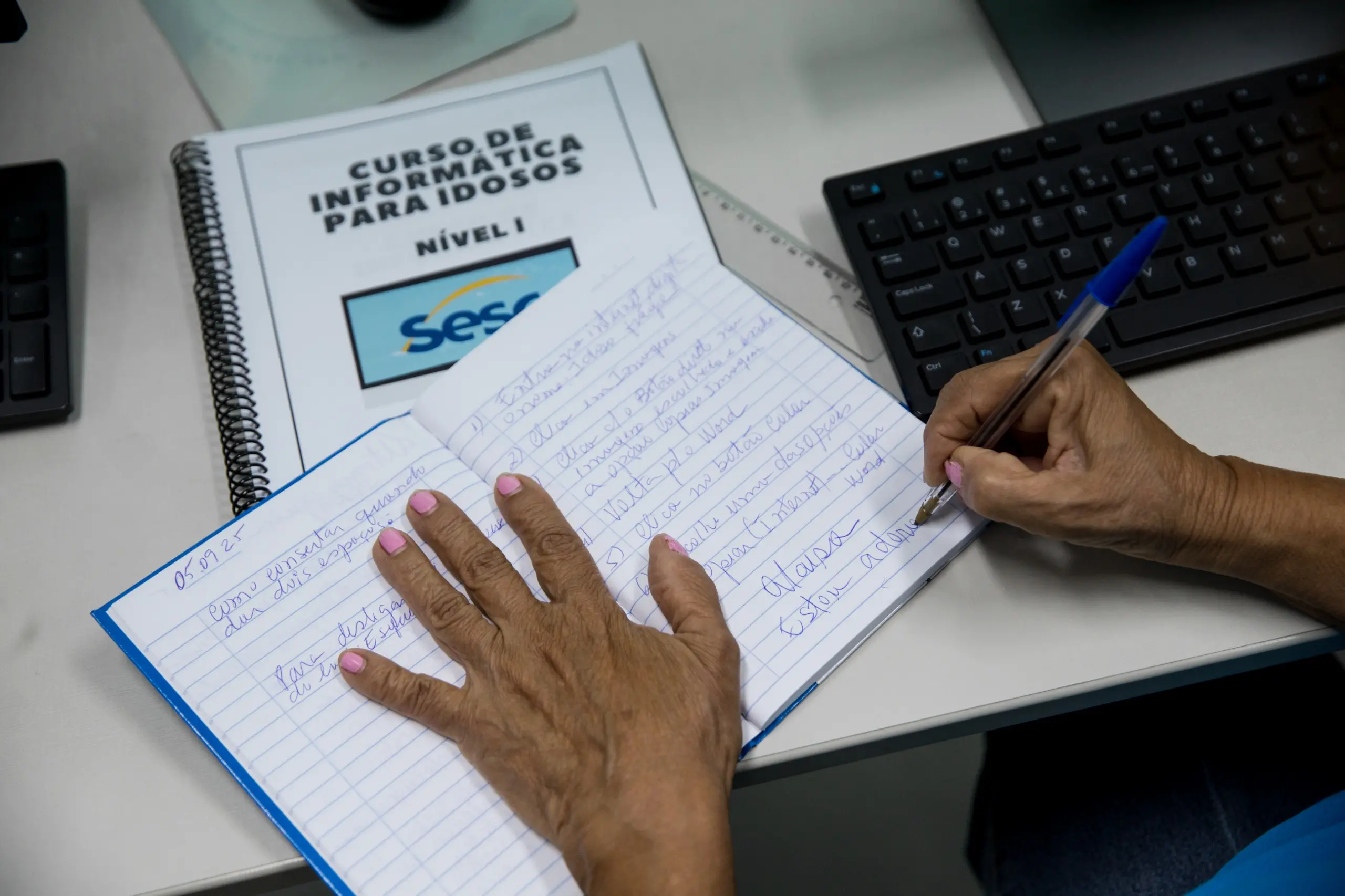 Imagem mostra mãos escrevendo em um caderno. Embaixo do caderno, há uma apostila com uma capa intitulada 