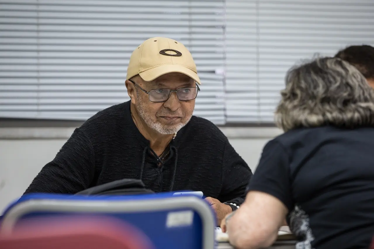 Homem de boné olhado para mulher de cabelo curto. Um de frente para o outro em mesa escolar.
